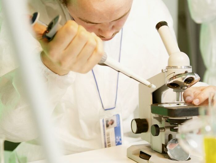 Scientist wearing a lab coat uses a microscope and pipette for research.